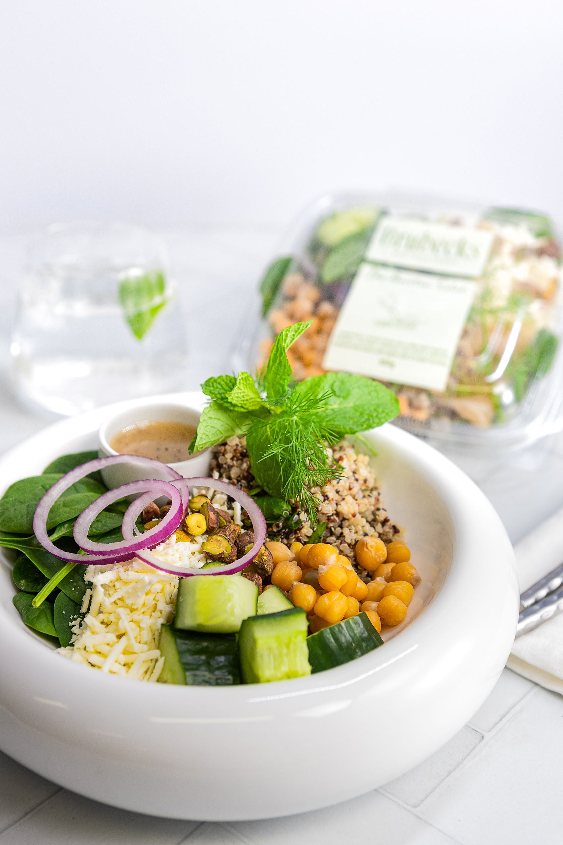 White bowl with salad ingredients including chickpeas, cucumbers, and red onions on a white surface.