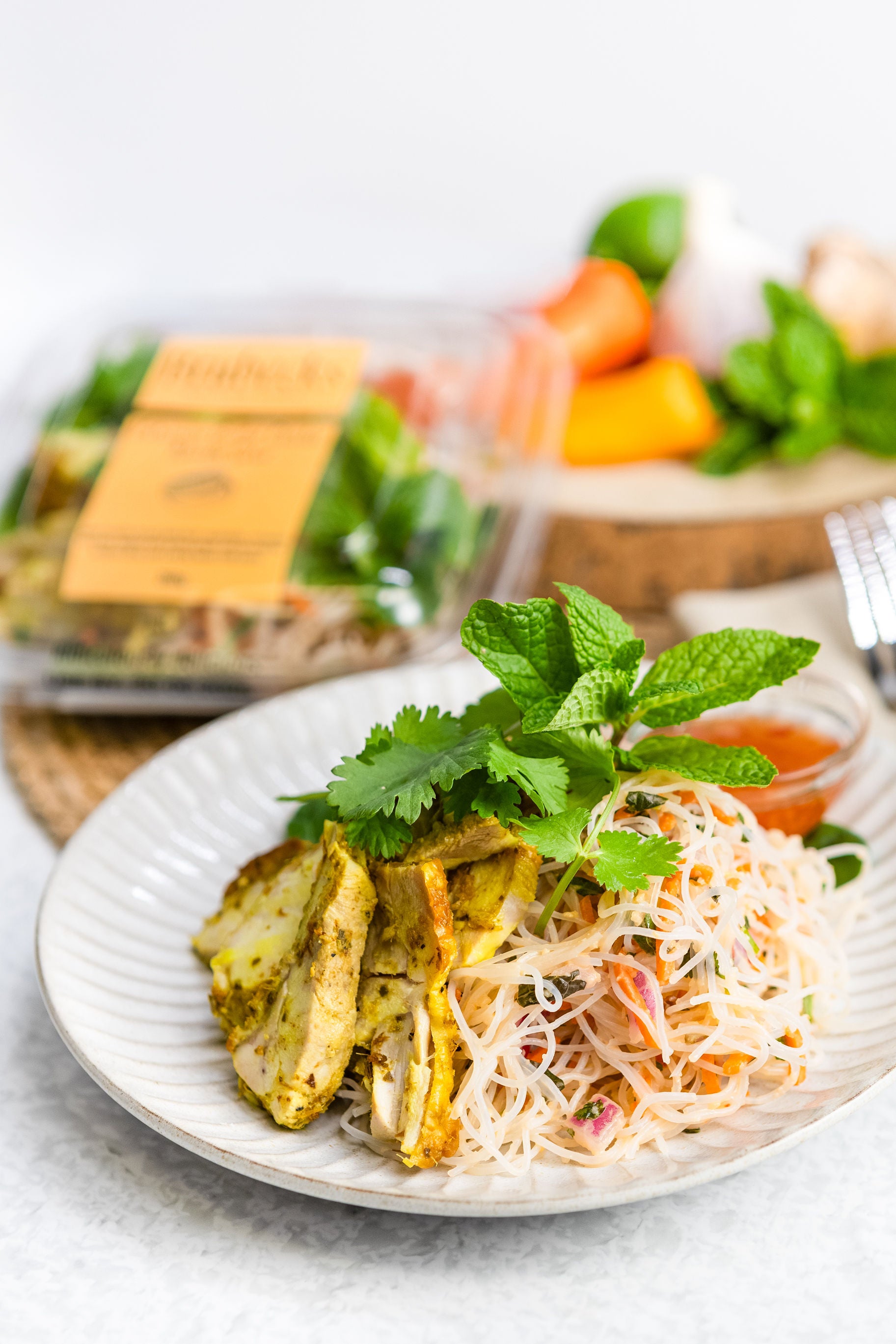 Plated dish with noodles, vegetables, and herbs on a white plate with a blurred background of ingredients.