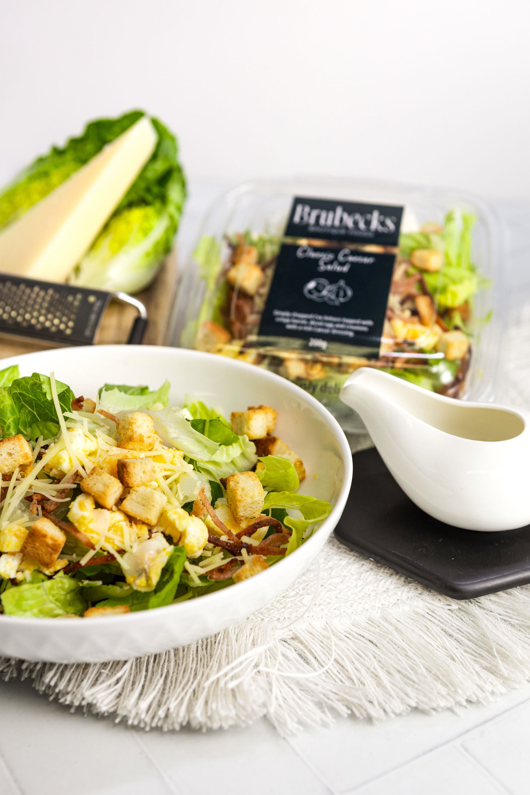 Caesar salad with croutons in a white bowl, surrounded by ingredients and a package on a white surface.