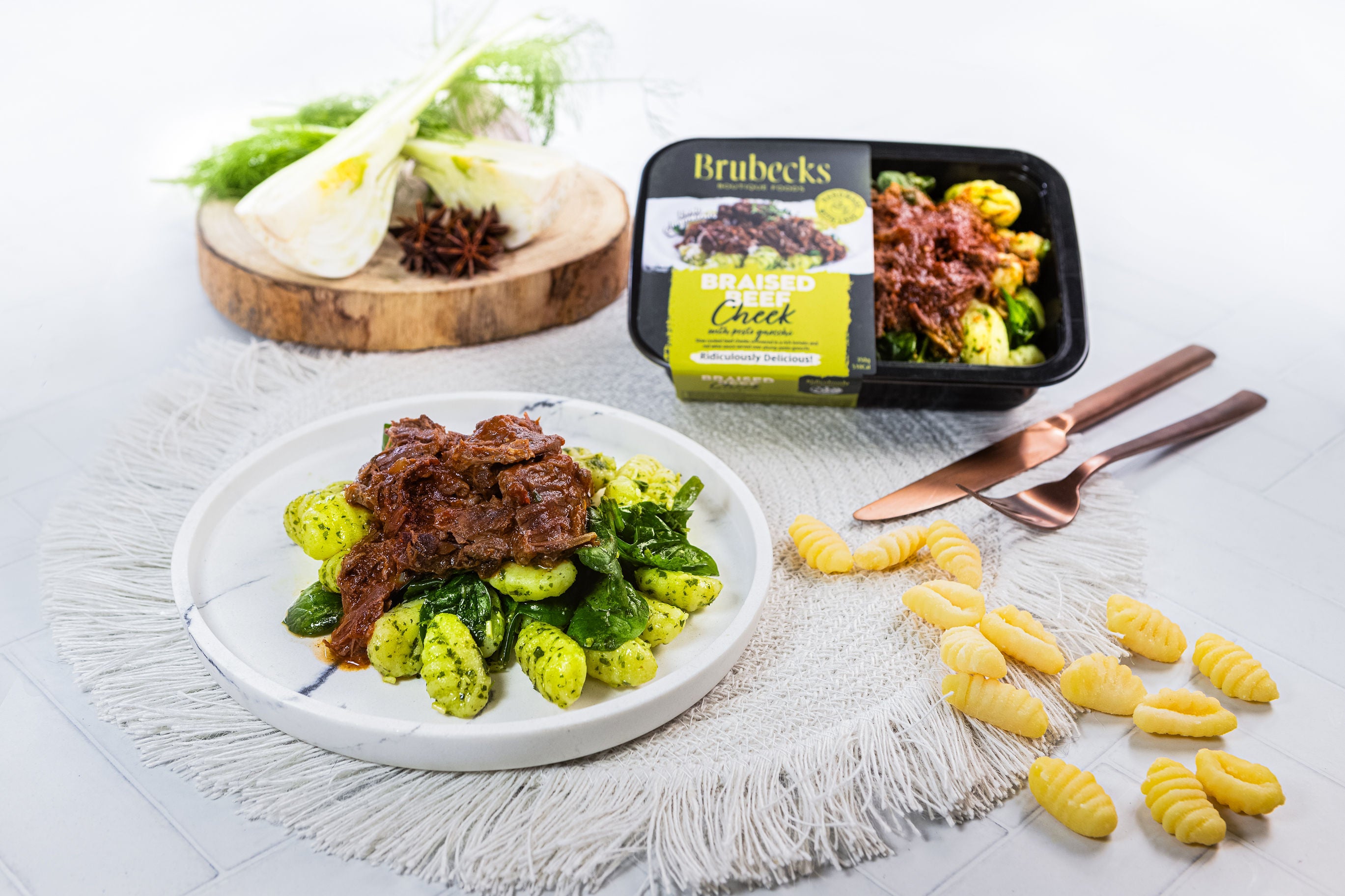 Plated dish of braised cheek with greens next to a container of Brubecks food product on a white surface.