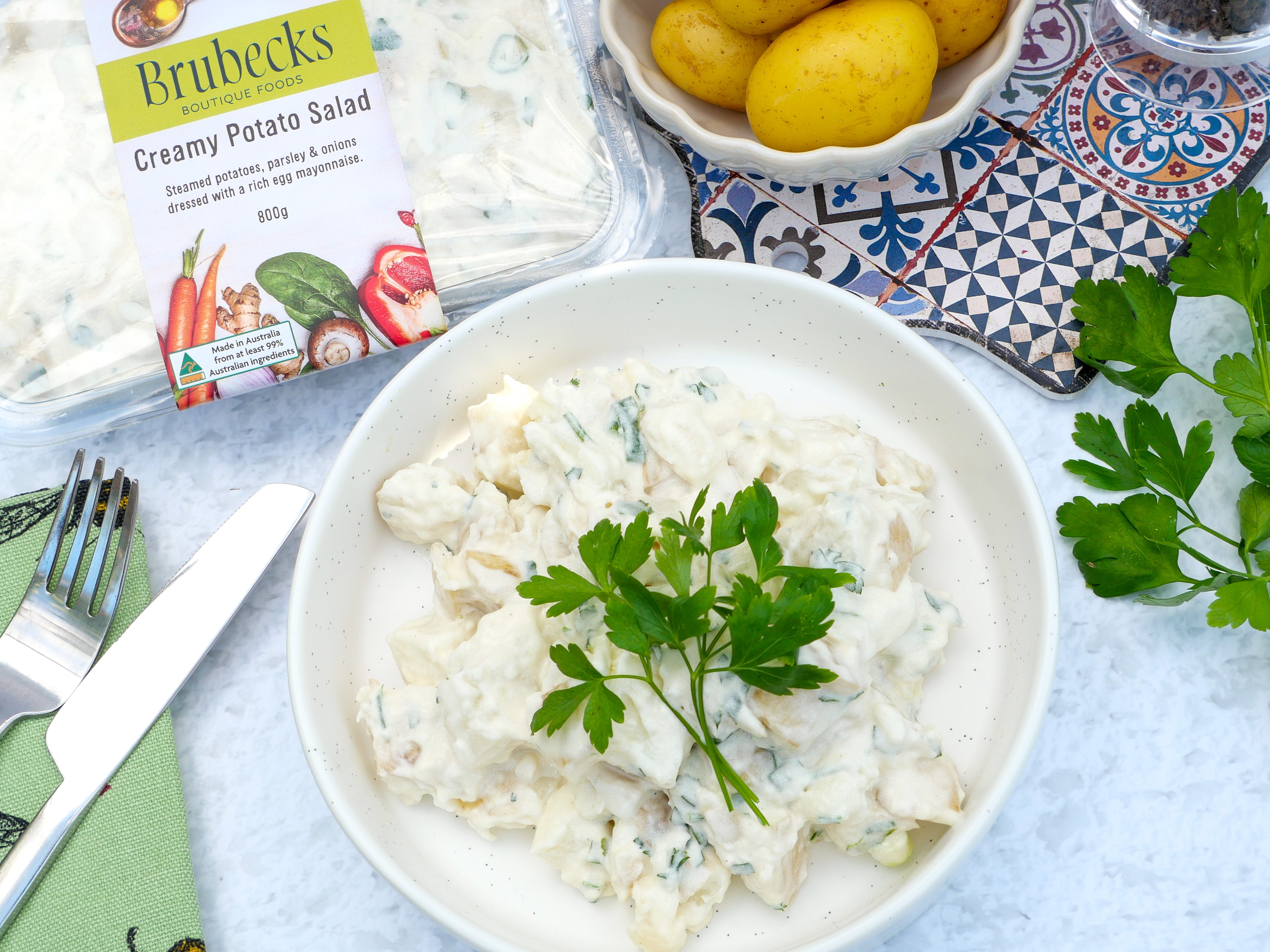 White bowl of creamy potato salad with parsley, Brubeck's creamy potato salad packaging, and potatoes on a patterned napkin.