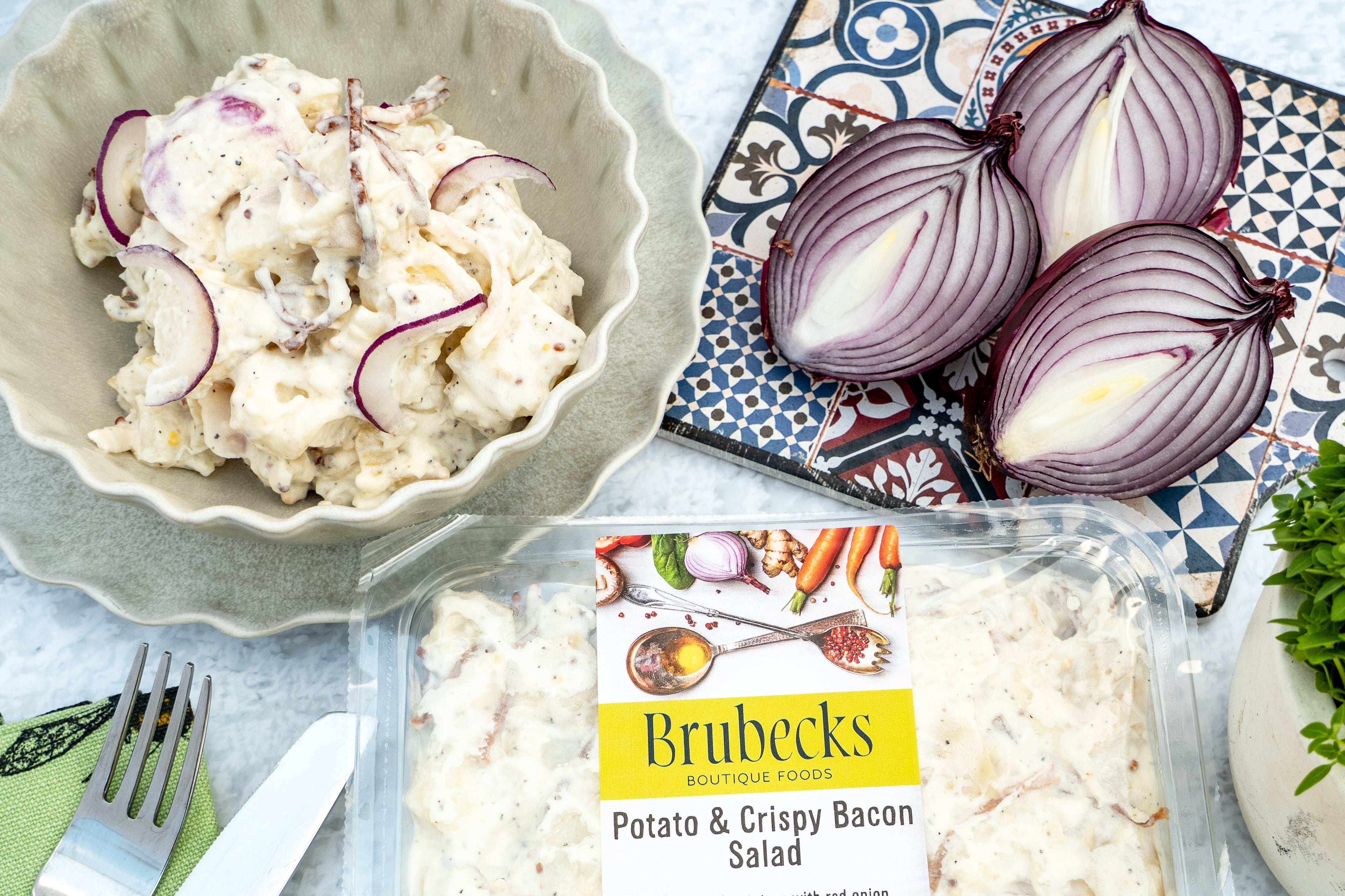 Brubecks Potato & Crispy Bacon Salad packaging with a bowl of salad and sliced red onions on a patterned tablecloth.