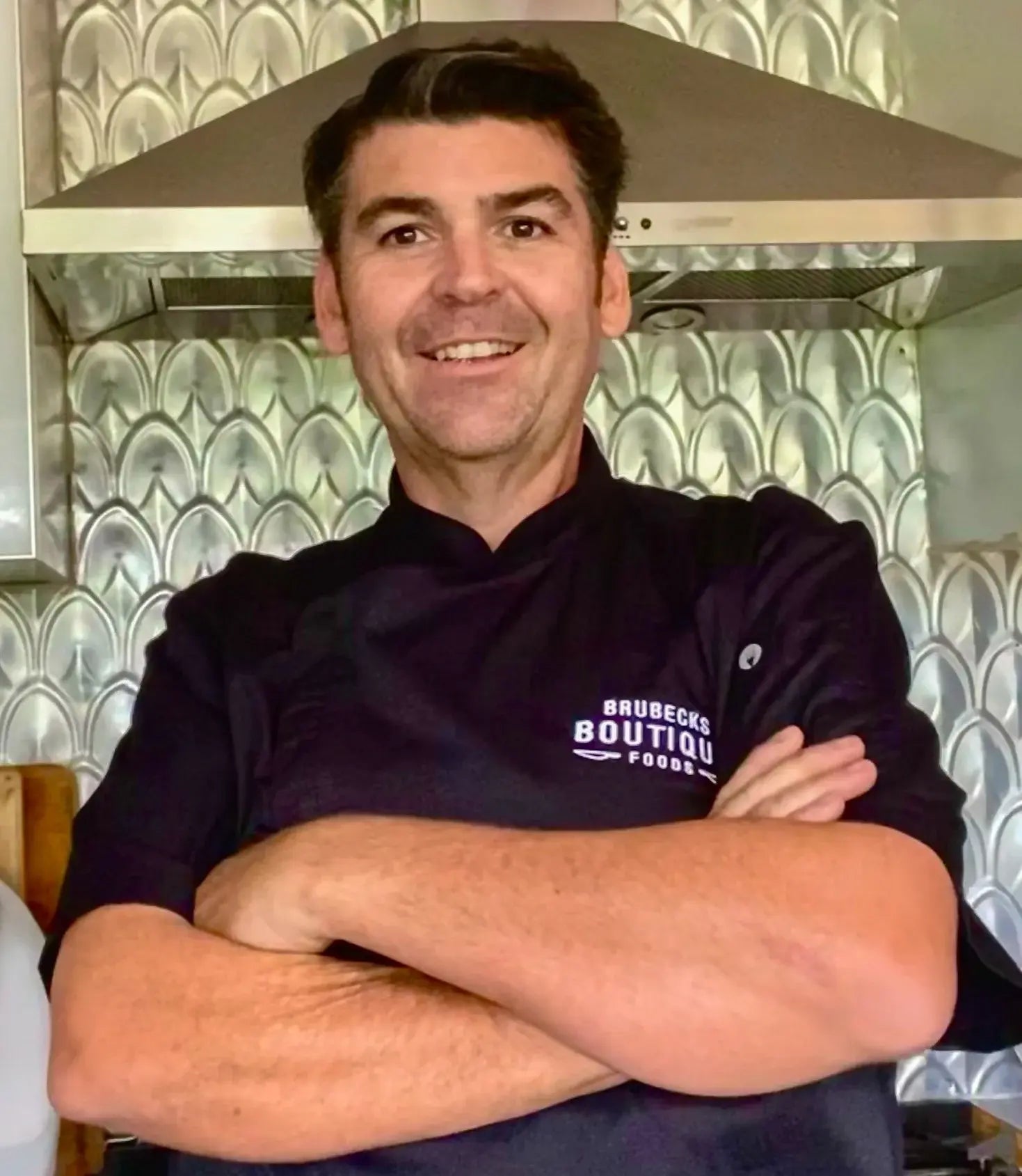 Chef Jamie Whitlock wearing a black chef's coat with 'Brubecks Boutique Foods' logo in a kitchen setting.