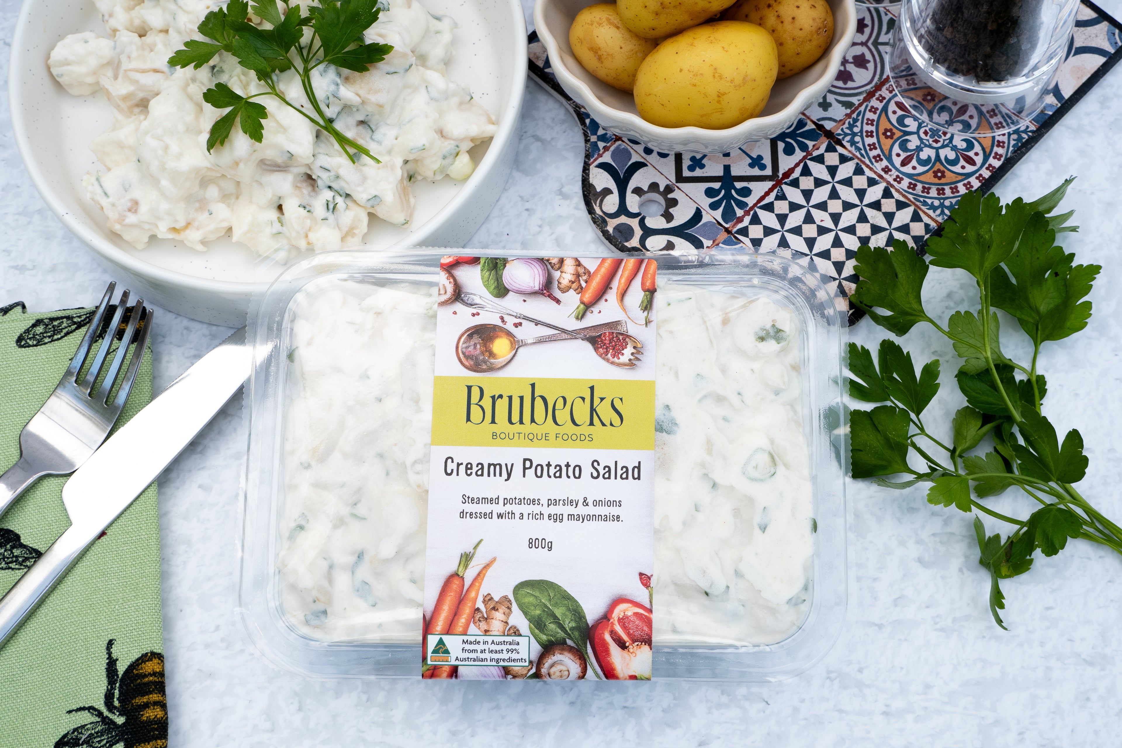 Packaged creamy potato salad with Brubecks branding on a table with a bowl of salad and potatoes.