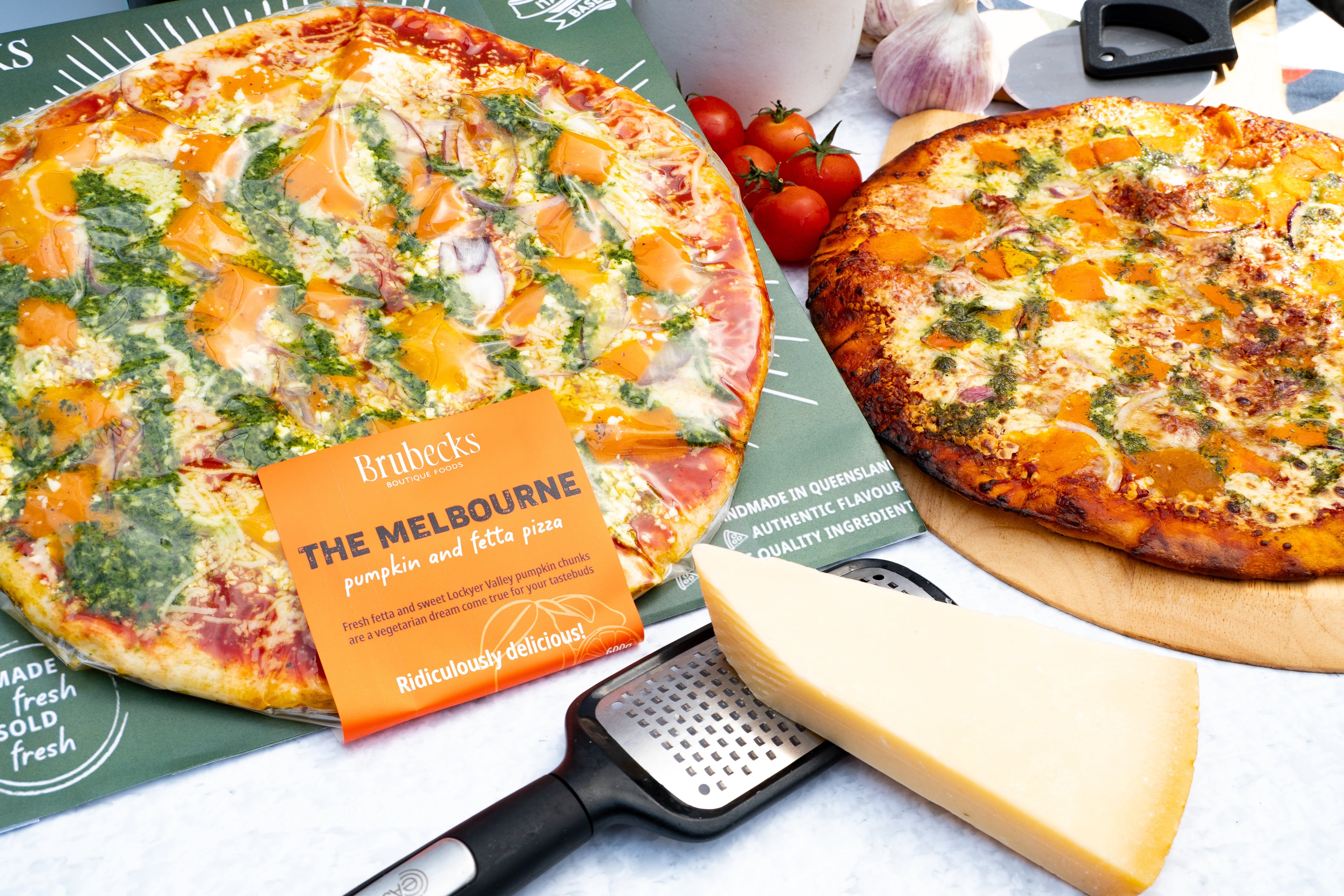 Two pizzas on a table with a grater and cheese block, featuring Brubeck's branding.