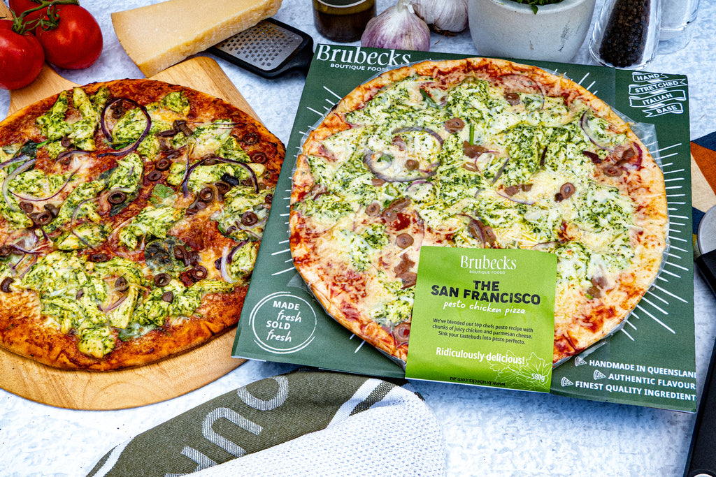 Two Brubecks San Francisco pizzas on a table with one pizza on a wooden board and the other in a branded box. 