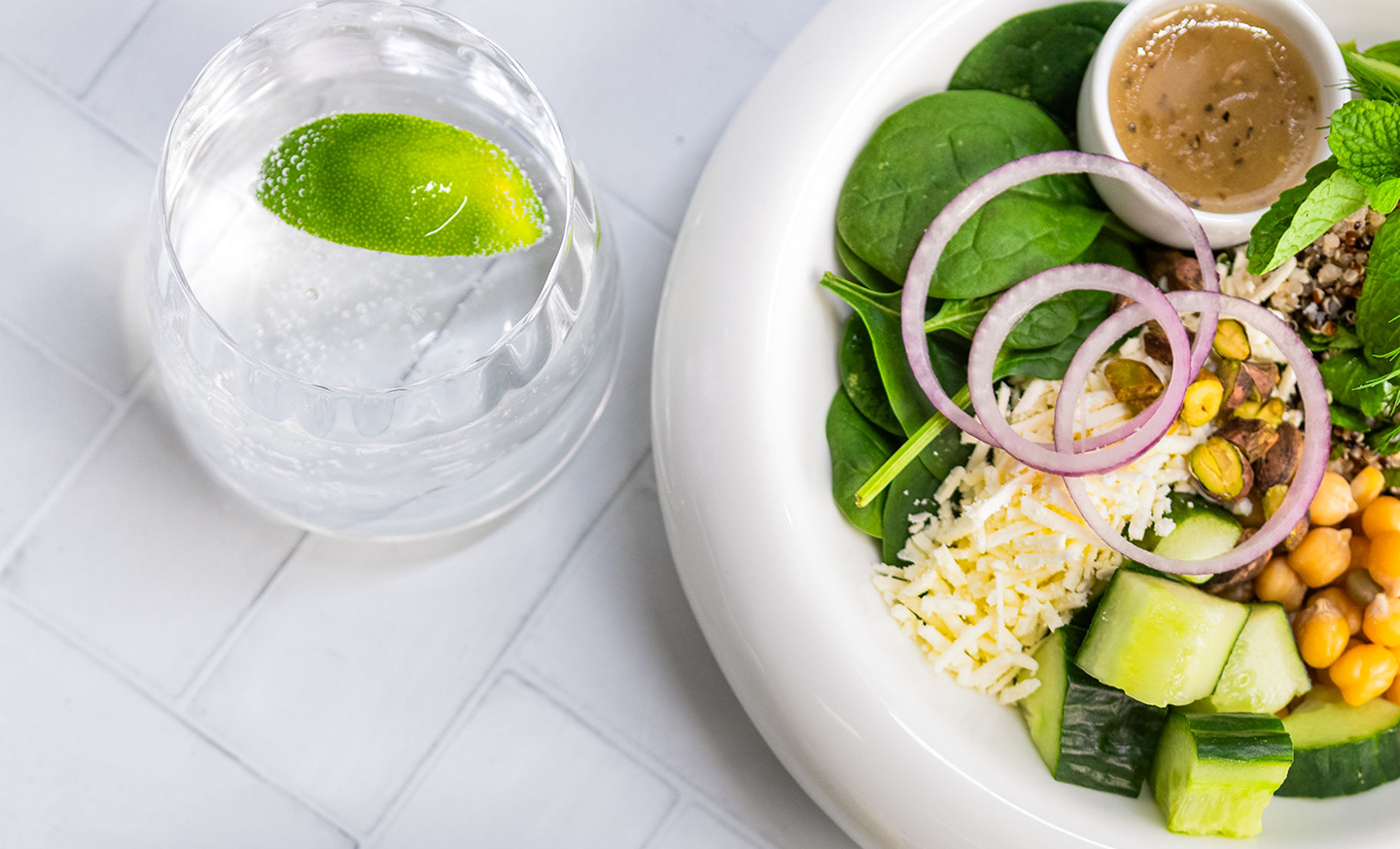 Brubecks salad with greens, cucumbers, chickpeas, and a small bowl of dressing on a white surface.