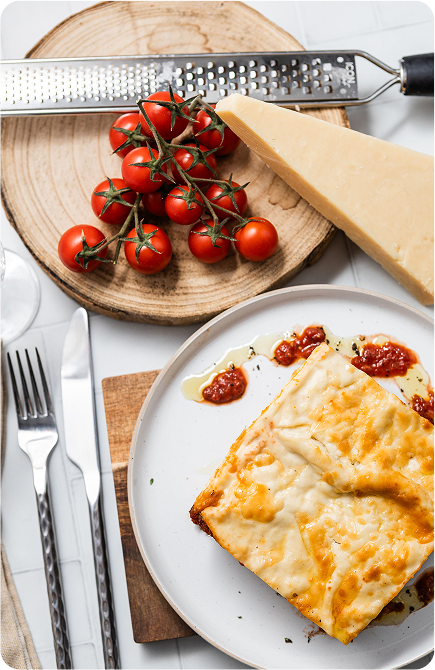Brubecks lasagne with tomato sauce on a plate, surrounded by tomatoes, a grater, and a block of cheese.
