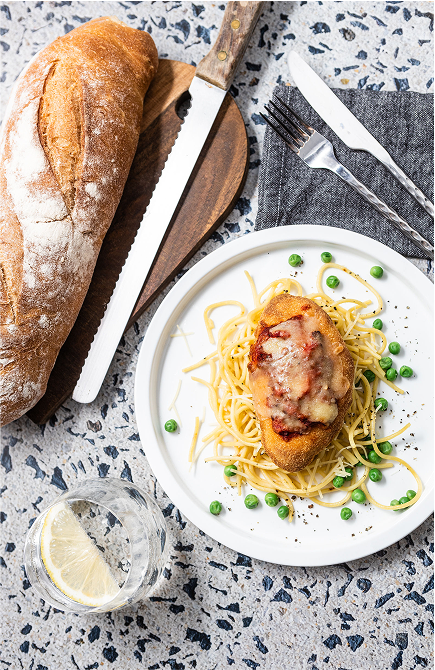 Plate of Brubecks spaghetti with a meatball, bread, knife, fork, and glass on a speckled surface